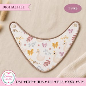 May include: A white triangular baby bib featuring a repeating pattern of swans wearing gold crowns, yellow bows, pink bows, and small floral sprigs. The bib has a brown stitched border and two beige snap buttons at the top corners for closure.