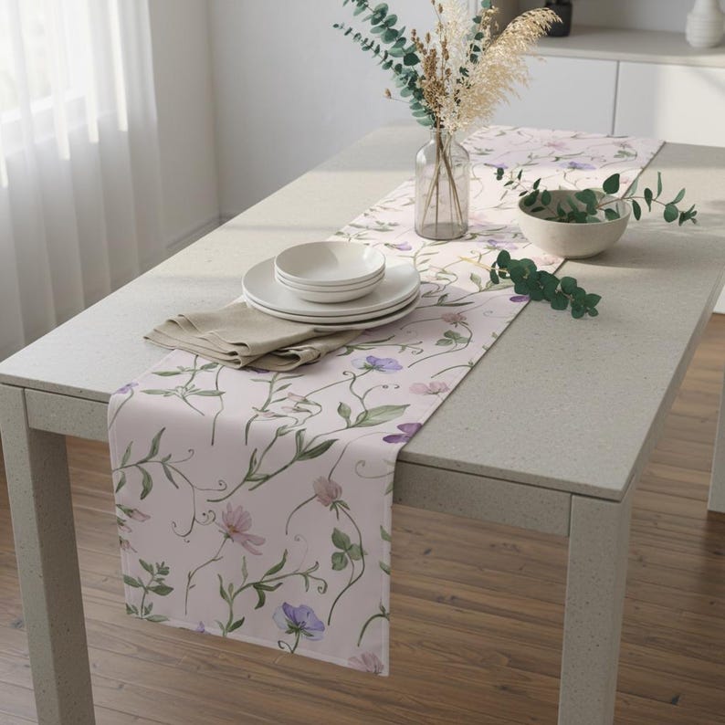 K&ouml;nnte beinhalten: Hellrosa Blumengedeckter Tischl&auml;ufer mit einem zarten Muster aus gr&uuml;nen Ranken und lila und rosa Bl&uuml;ten. Der L&auml;ufer liegt auf einem hellgrauen Tisch, mit wei&szlig;en Tellern, Schalen und einer beigen Serviette. Eine Vase mit Trockenblumen und Gr&uuml;nzeug.