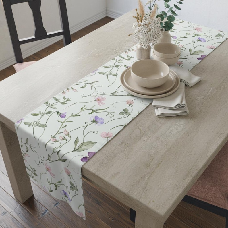 K&ouml;nnte beinhalten: Ein floraler Tischl&auml;ufer mit hellem Hintergrund und einem Muster aus rosa und lila Blumen mit gr&uuml;nen Bl&auml;ttern. Der L&auml;ufer liegt auf einem hellen Holztisch mit beigen Tellern, Schalen und Servietten.