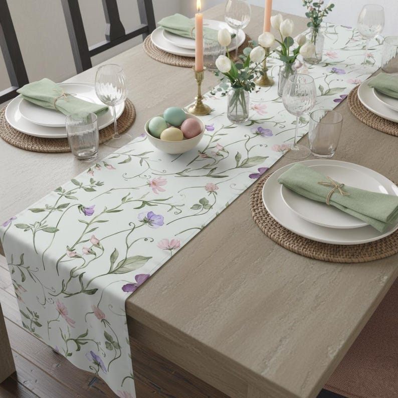 May include: A floral table runner featuring a light background and a pattern of pink and purple flowers and green vines. The table is set with white plates, green napkins, and decorative elements like candles and a bowl of pastel-coloured eggs.