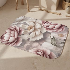 May include: Rectangular bath mat featuring a floral design with large, detailed flowers in shades of pink, white, and gray. The mat has a soft, plush appearance, suitable for a bathroom setting.