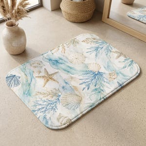 Op de afbeelding: Een rechthoekige badmat met een aquarelprint van diverse schelpen, zeesterren en blauwe koraaltakken op een witte achtergrond. Het ontwerp bevat zachte blauwe en beige tinten, met afgeronde hoeken en een afgewerkte rand.