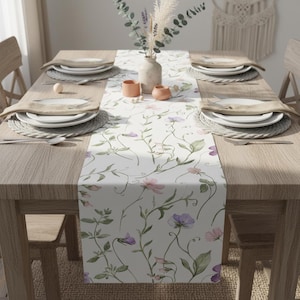 K&ouml;nnte beinhalten: Ein Holztisch, gedeckt f&uuml;r eine Mahlzeit, mit einem floralen Tischl&auml;ufer mit rosa und lila Blumen. Der Tisch ist mit Tellern, Tischsets und Besteck gedeckt. Eine Vase mit Trockenblumen und kleine Keramikschalen stehen auf dem Tisch.