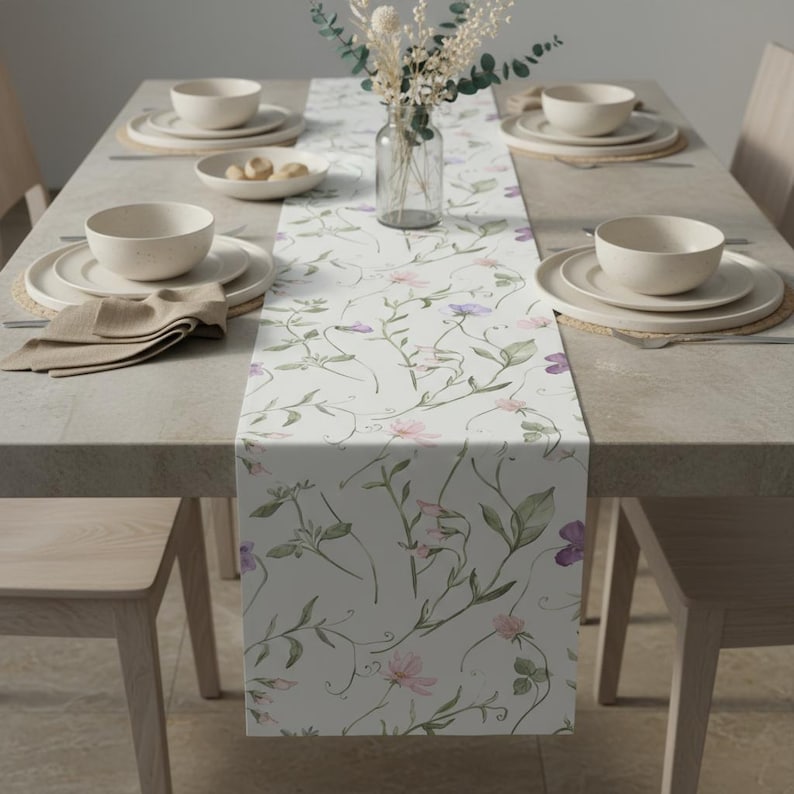 K&ouml;nnte beinhalten: Ein gedeckter Esstisch mit einem floralen Tischl&auml;ufer. Der L&auml;ufer hat einen wei&szlig;en Hintergrund mit rosa und lila Blumenmustern. Beige Schalen, Teller und Servietten sind auf dem Tisch platziert. Eine klare Vase mit getrockneten Blumen erg&auml;nzt die Dekoration.
