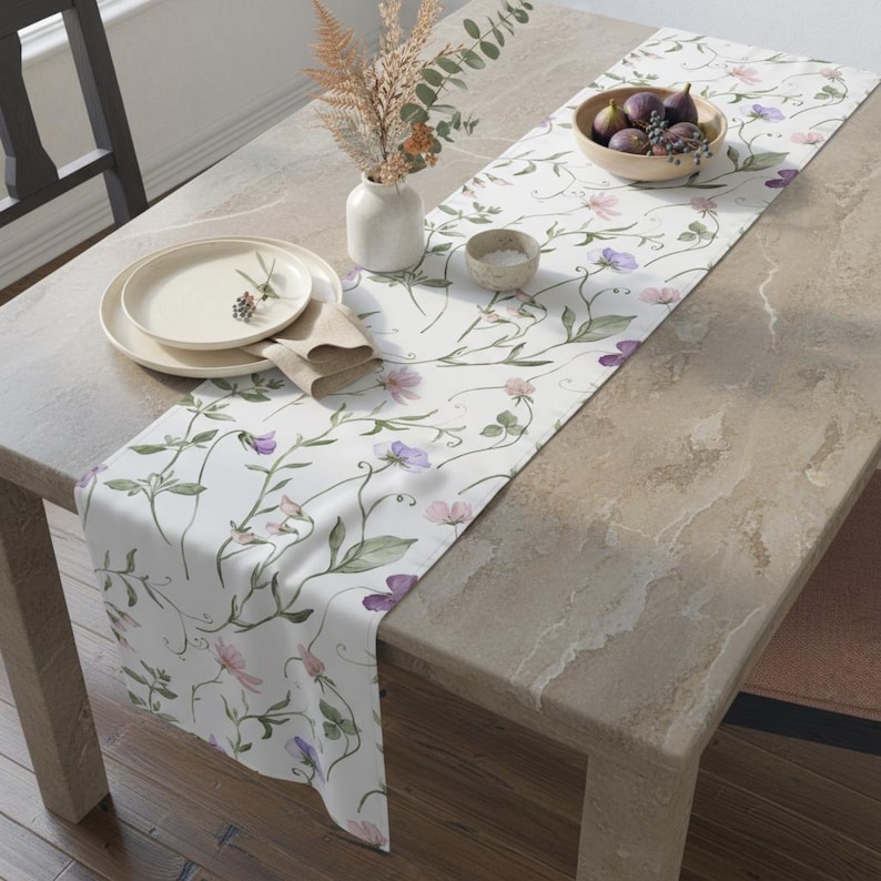 K&ouml;nnte beinhalten: Ein wei&szlig;er Tischl&auml;ufer mit einem Blumendesign mit lila und rosa Blumen und gr&uuml;nen Ranken. Der L&auml;ufer liegt auf einem hellen Tisch, zusammen mit einer Schale mit Feigen, Tellern und einer Vase mit getrockneten Blumen. Ein dekoratives St&uuml;ck f&uuml;r das Esszimmer.