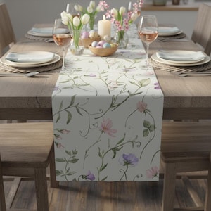 May include: A cream-coloured table runner featuring a floral design of pink and purple flowers and green leaves. The runner is placed on a wooden table, complemented by place settings, wine glasses, and decorations.