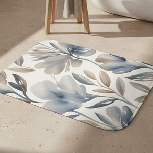 May include: Rectangular bath mat featuring a watercolor floral design. The mat has a white background with blue, gray, and brown floral and leaf patterns. The mat is placed on a light-colored floor.