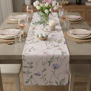 K&ouml;nnte beinhalten: Ein Esstisch, der f&uuml;r eine Mahlzeit gedeckt ist, mit einem hellrosa Blumengedeck mit lila und gr&uuml;nen Akzenten. Der Tisch ist mit Tellern, Gl&auml;sern und Besteck gedeckt. Eine Vase mit wei&szlig;en und rosa Blumen und eine Kerze sind ebenfalls vorhanden.