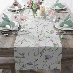 K&ouml;nnte beinhalten: Ein gedeckter Tisch mit einem floralen Tischl&auml;ufer mit rosa und lila Blumen und gr&uuml;nen Ranken. Der Tisch ist mit Tellern, Gl&auml;sern und hasenf&ouml;rmigen Serviettenhaltern gedeckt. Eine Vase mit Blumen und eine brennende Kerze stehen ebenfalls auf dem Tisch.