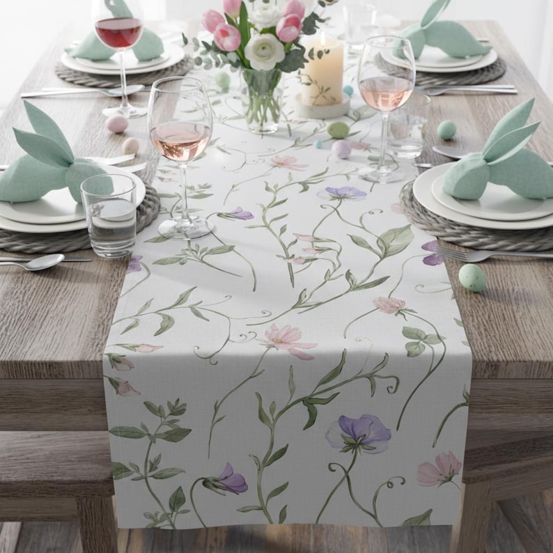 K&ouml;nnte beinhalten: Ein gedeckter Tisch mit einem floralen Tischl&auml;ufer mit rosa und lila Blumen und gr&uuml;nen Ranken. Der Tisch ist mit Tellern, Gl&auml;sern und hasenf&ouml;rmigen Serviettenhaltern gedeckt. Eine Vase mit Blumen und eine brennende Kerze stehen ebenfalls auf dem Tisch.