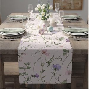 Peut inclure: Chemin de table floral avec un fond rose clair et un motif aquarelle de fleurs violettes et roses et de vignes vertes. La table est dress&eacute;e avec des assiettes blanches, des serviettes vertes et des verres &agrave; vin. Un vase de tulipes blanches et des &oelig;ufs d&eacute;coratifs sont sur la table.