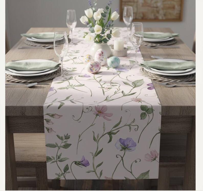 Peut inclure: Chemin de table floral avec un fond rose clair et un motif aquarelle de fleurs violettes et roses et de vignes vertes. La table est dress&eacute;e avec des assiettes blanches, des serviettes vertes et des verres &agrave; vin. Un vase de tulipes blanches et des &oelig;ufs d&eacute;coratifs sont sur la table.