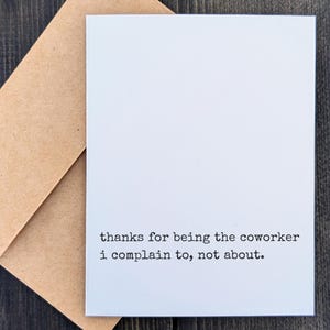 May include: A white greeting card with the text "thanks for being the coworker I complain to, not about." is paired with a brown envelope. The card and envelope are on a dark wood surface.