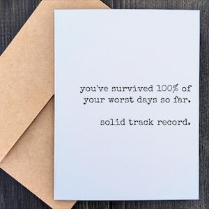 May include: A white greeting card with the text "you've survived 100% of your worst days so far. solid track record." is paired with a brown envelope. The card and envelope are on a dark wood surface.