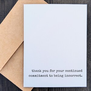 May include: A white greeting card with the text "thank you for your continued commitment to being incorrect." is placed on top of a brown envelope. The card and envelope are on a dark wooden surface.