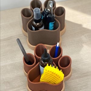 Könnte beinhalten: Zwei braune, pfotenförmige Organizer mit mehreren Fächern. Einer enthält Pflegeutensilien und Sprühflaschen, der andere eine Feile, eine Flasche, einen blauen Stift und eine gelbe Bürste. Die Organizer stehen auf einer hellen Oberfläche.