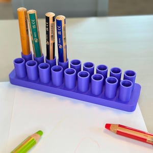 May include: A lilac pencil holder with 18 cylindrical holes, containing four pencils of different colours. Two coloured pencils lie on the white paper in the foreground. The pencils are orange, green, black, and blue.