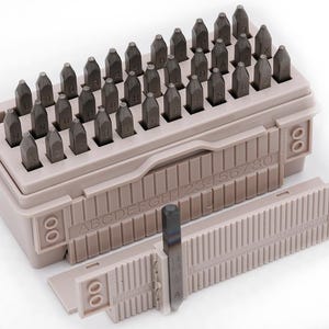 May include: A set of metal letter and number stamps in a beige plastic storage case. The stamps are arranged in rows and columns, with a sliding guide. The case is labeled with the alphabet and numbers.
