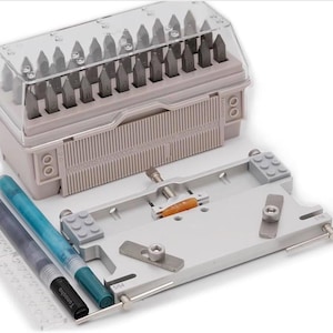 May include: A metal stamping kit with a clear plastic lid, containing numerous metal stamps. The kit includes a grey base with various components, and two pens. The pens are black and teal. The kit is designed for metalworking.