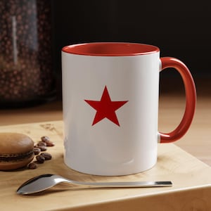 May include: White ceramic mug with a red interior, handle, and a large red star on the front. The mug sits on a wooden cutting board with a spoon, coffee beans, and a macaron.