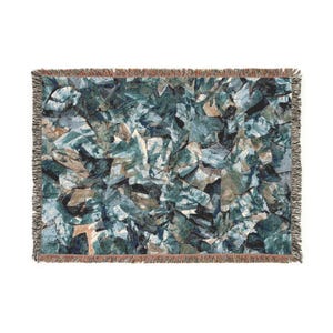 May include: A rectangular throw blanket with a geometric pattern in shades of blue, green, and brown. The design resembles abstract shapes or fractured stone. The blanket has a fringed edge.