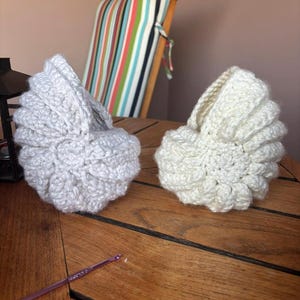 May include: Two handmade crochet items, one light grey and one off-white, sit on a wooden surface. The items have a rounded, shell-like shape. A purple crochet hook lies nearby. A chair with a striped cushion is in the background.
