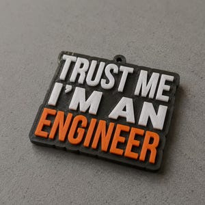 May include: A black and grey rectangular keyring with the text "TRUST ME I'M AN ENGINEER" in white and orange lettering. The keyring has a small hole at the top for attachment.