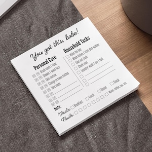 May include: A white notepad with black text reading "You got this, babe!" and lists for personal care and household tasks. The notepad includes checkboxes for meals and fluids. The notepad is on a gray surface.