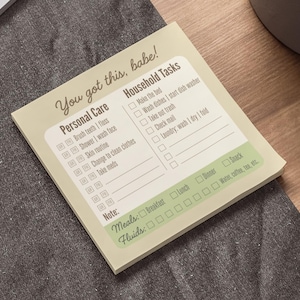 May include: A notepad with a light green background and text that reads "You got this, babe!". The notepad is divided into sections for personal care and household tasks, with checkboxes for each item. The notepad is on a gray surface.