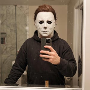 Könnte beinhalten: Eine weiße Michael Myers Maske mit dunklen Augenhöhlen und braunem Haar, getragen mit einem schlichten schwarzen Kapuzenpullover. Die Maske zeigt einen neutralen Ausdruck mit definierten Gesichtszügen. Ein Smartphone wird für das Spiegelbild gehalten.