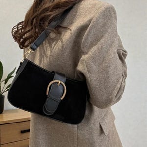 May include: A black suede shoulder bag with a rectangular shape. The bag features a black leather strap and a gold-colored buckle closure. The bag is worn over a brown blazer.