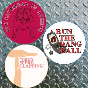 May include: Four round buttons with text and graphics. One button is pink with a bulldog and the text "It's Saturday in Athens". Another button has a phone with a football and the text "Run the Dang Ball". The last button has a hand gesture and the text "They Were Clapping".