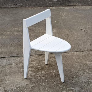 May include: A white wooden chair with a triangular seat and a backrest. The chair has three legs and is painted white. The chair is designed with a minimalist aesthetic.