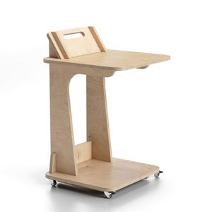 May include: A light-coloured wooden laptop desk on wheels. The desk has a rectangular top with a raised back section and a handle. The desk is supported by a vertical frame with a lower shelf and four small wheels.