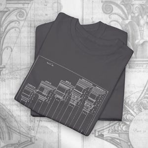 May include: Two charcoal gray t-shirts are stacked. The top shirt features a white architectural blueprint design with the text "TUSCAN, DORIC, IONIC, CORINTHIAN, COMPOSITE". The design includes detailed illustrations of classical columns.