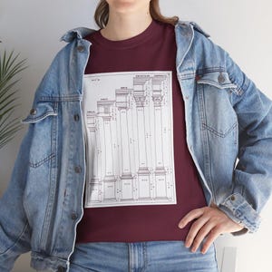 May include: A maroon t-shirt featuring a detailed architectural diagram of various column styles, including Tuscan, Doric, Ionic, Corinthian, and Composite. The diagram is in black and white. The person is wearing a light blue denim jacket.