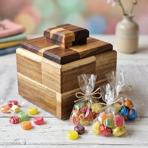 Può includere: Una scatola quadrata in legno con un coperchio a scacchiera e un pomello quadrato abbinato. La scatola è accompagnata da tre sacchetti di plastica trasparente pieni di caramelle colorate, legati con spago, e caramelle sparse vicino.