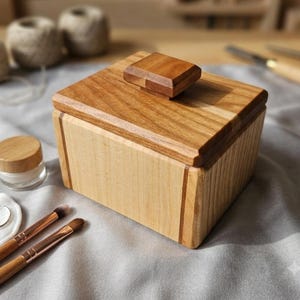 Può includere: Una scatola rettangolare in legno con un corpo di colore chiaro e un coperchio in legno lucidato più scuro dotato di una maniglia centrale quadrata. La scatola presenta dettagli scanalati verticali sui lati e una superficie liscia.