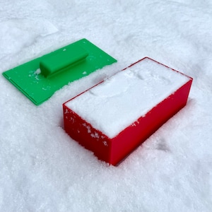 Può includere: Un raschietto di plastica verde brillante e una scatola rettangolare rossa, entrambi coperti di neve, poggiano su una superficie innevata. Il raschietto ha un manico e una lama piatta. La scatola è di colore rosso pieno.