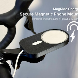 Puede incluir: Soporte magnético negro para teléfono, fijado al manillar de una bicicleta. El soporte tiene una superficie de carga blanca y lleva la inscripción "MagRide Charge". Compatible con MagSafe V1 (15W) y V2 (25W).