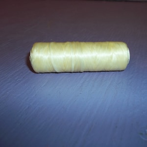 May include: A cylindrical spool of pale yellow thread. The thread is tightly wound, creating a smooth, textured surface. The spool is resting on a purple surface.