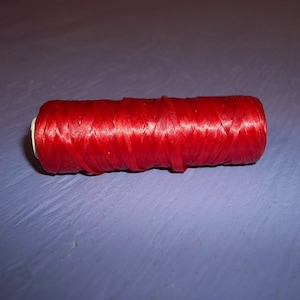 May include: A spool of bright red thread, tightly wound and cylindrical in shape. The thread has a slightly shiny texture, and the spool has white ends. This item is likely used for sewing or crafting.