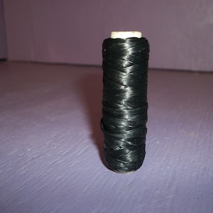 May include: A spool of black thread, tightly wound around a cylindrical core. The thread has a slight sheen, reflecting light. The spool is standing upright against a purple background. This thread is likely used for sewing or crafting.