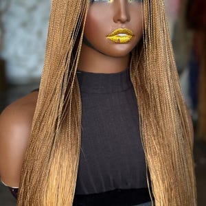 May include: A mannequin head with a long, braided wig in shades of light brown and gold. The wig is styled with a center part and falls to the mid-chest. The mannequin has dark skin, gold glitter lips, and gold eyeshadow. The mannequin is wearing a black top.