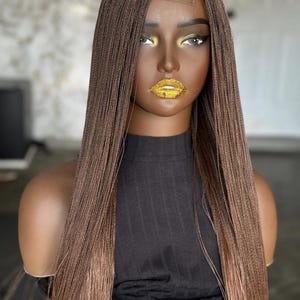 May include: A brown wig with long, micro braids styled on a mannequin head. The wig has a lace front and a dark brown to light brown ombre effect. The mannequin is wearing a black top and has gold glitter lips.