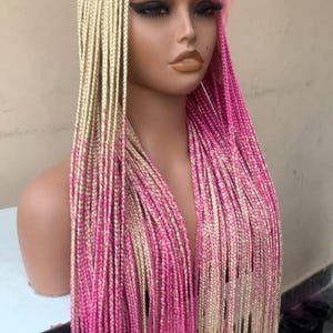 May include: A blonde and pink braided wig on a mannequin head. The wig features long, straight braids with a gradient effect, transitioning from blonde to vibrant pink. The wig is styled on a mannequin with a neutral skin tone.