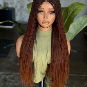 May include: A mannequin head displaying a long, braided wig with a natural-looking hairline. The wig transitions from a dark brown at the roots to a lighter, reddish-brown at the ends. The mannequin wears a light green turtleneck.