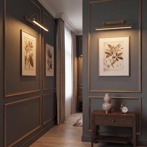 May include: A hallway with dark gray walls featuring gold-toned molding, illuminated by two horizontal brass wall lamps. The space includes framed botanical prints, a wooden console table with a beige ceramic vase, and a small analog alarm clock.