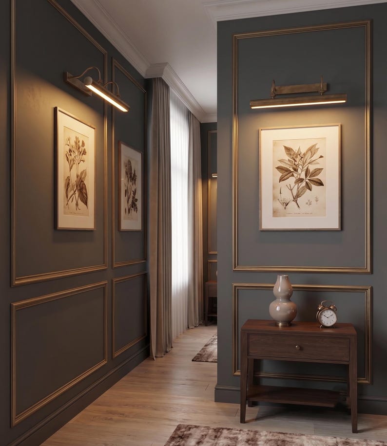 May include: A hallway with dark gray walls featuring gold-toned molding, illuminated by two horizontal brass wall lamps. The space includes framed botanical prints, a wooden console table with a beige ceramic vase, and a small analog alarm clock.