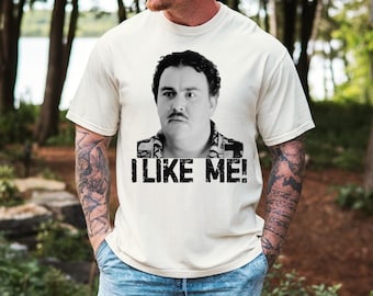 May include: A cream-colored short-sleeve t-shirt featuring a black and white photographic print of a man with a mustache and curly hair. Below the image, the text "I LIKE ME!" is printed in a bold, distressed black sans-serif font.